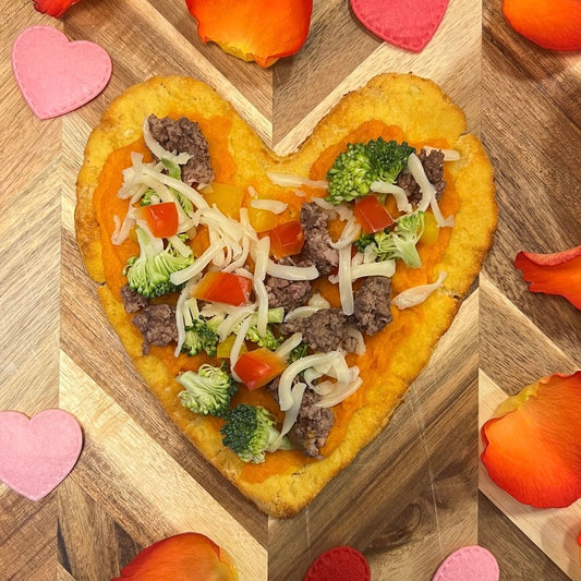 A heart-shaped, dog-friendly pizza made from a cauliflower dough topped with various vegetables, cheese, and ground beef.