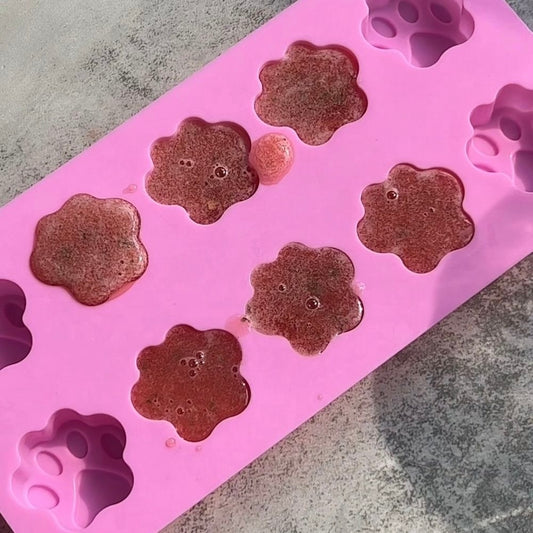 A pink mold of 10 paw print-shaped wells sits on a gray counter. The middle 6 wells are filled with a reddish pupsicle mix..