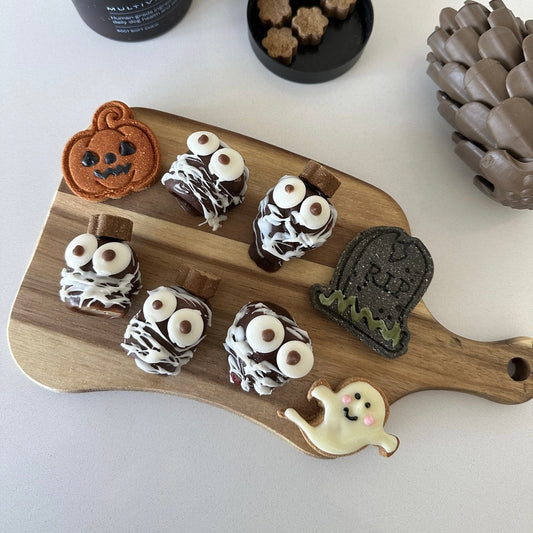 A wooden board with 5 banana carob ghost treats and three Halloween themed dog cookies. Two dog multivitamin jars and a pinecone are arranged next to the board.
