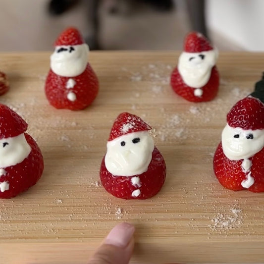 Five Santa-themed treats made from halves of strawberries sit on a wooden board. Dog-safe cookies and multivitamins decorate the board.