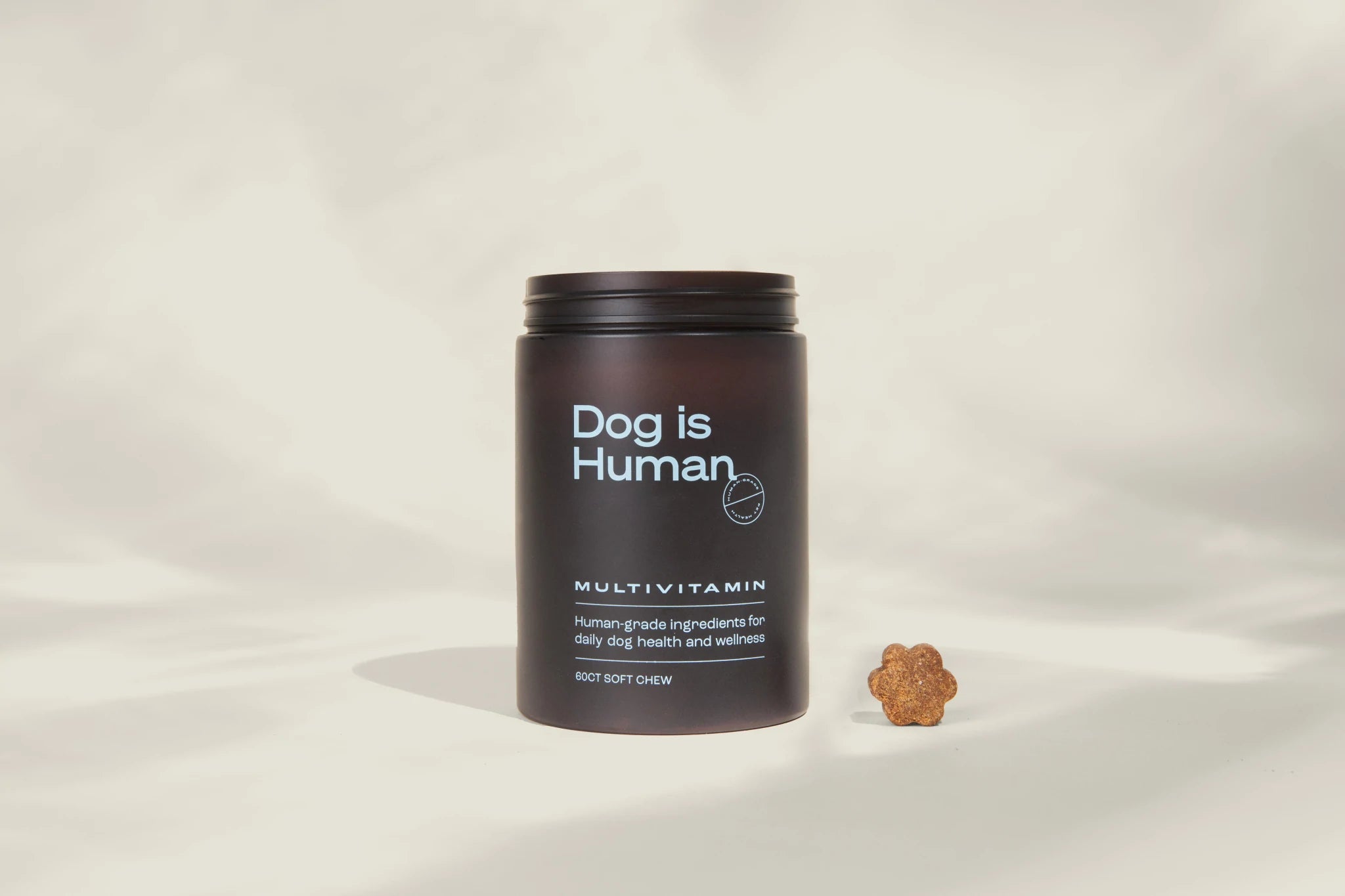 Front view of the Dog is Human multivitamin jar and one chew next to it against a beige, shadowy background.